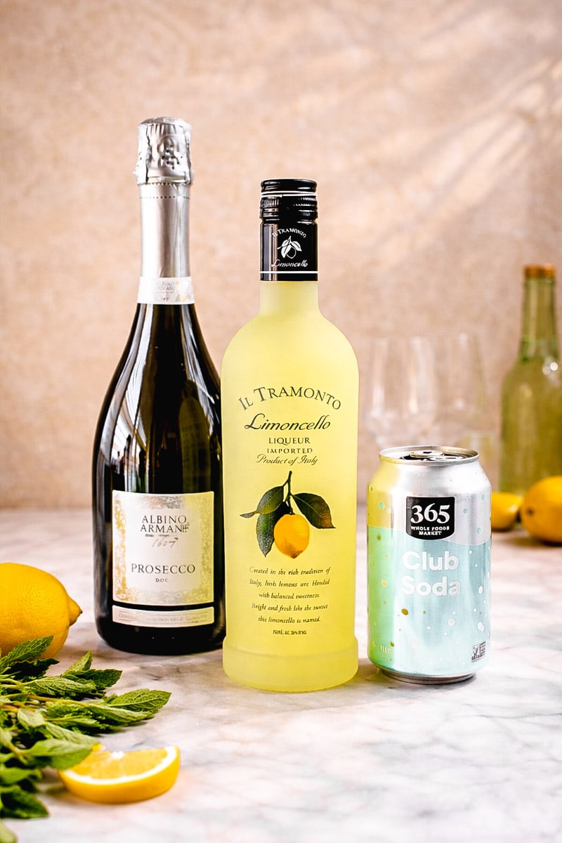 A bottle of Prosecco, a bottle of Il Tramonto Limoncello, and a can of 365 Club Soda are arranged on a marble surface with fresh lemons, mint, and empty glasses in the background.