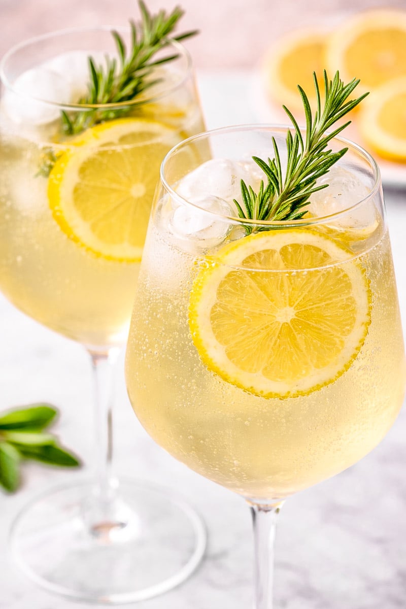 Two glasses of a light, bubbly drink with ice cubes, garnished with lemon slices and sprigs of rosemary; additional lemon slices and greenery are in the blurred background- limoncello spritz.