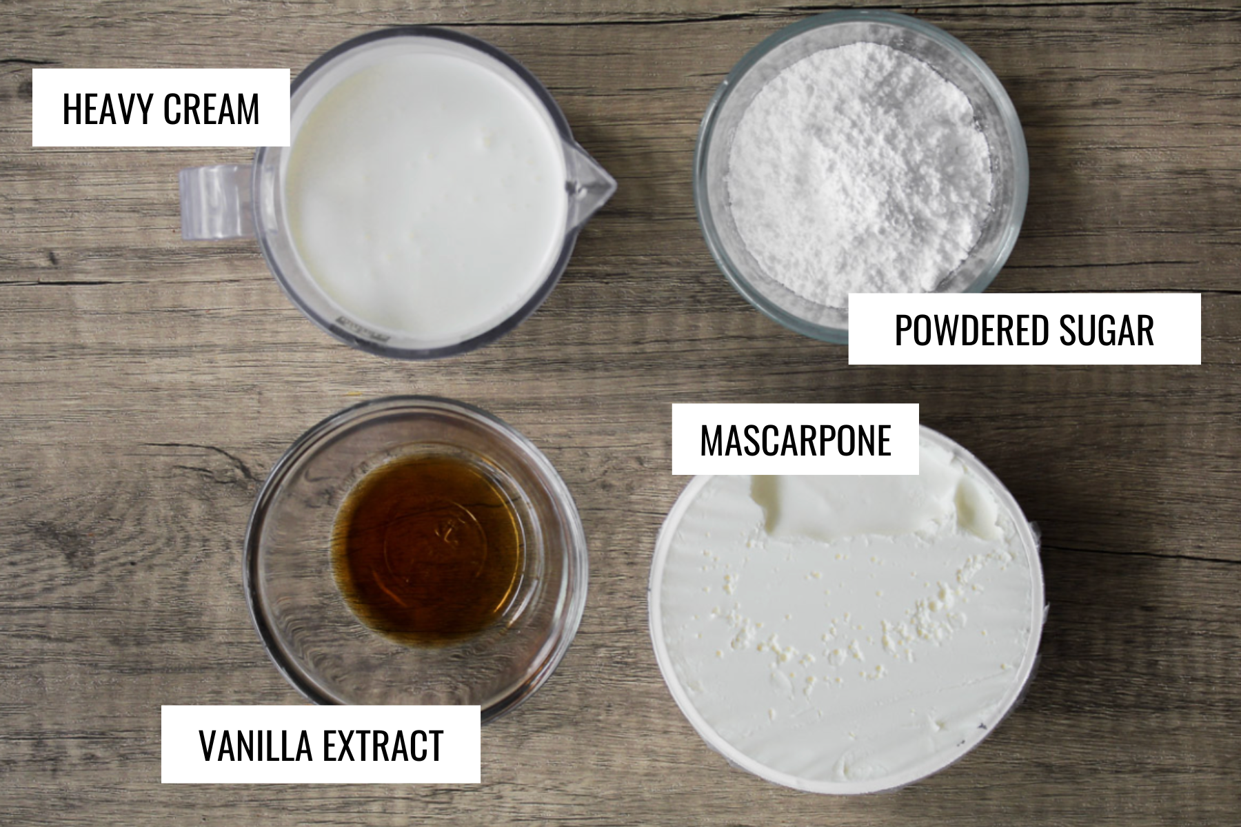Top-down view of four labeled ingredients for mascarpone cream on a wooden surface: a measuring cup of heavy cream, a bowl of powdered sugar, a bowl of mascarpone, and a small bowl of vanilla extract.
