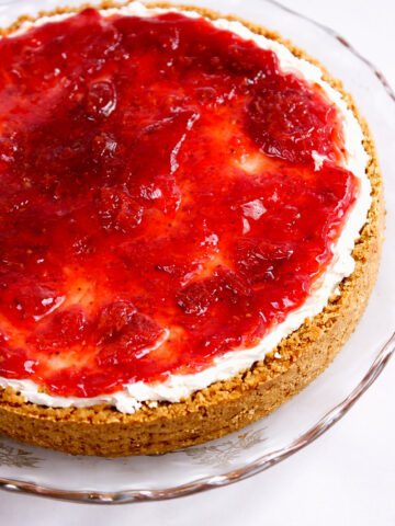 A cheesecake with a crumbly graham cracker crust, topped with a layer of creamy filling and a thick, glossy strawberry sauce, served on a clear glass plate.