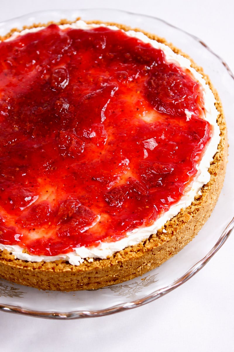 A cheesecake with a crumbly graham cracker crust, topped with a layer of creamy filling and a thick, glossy strawberry sauce, served on a clear glass plate.