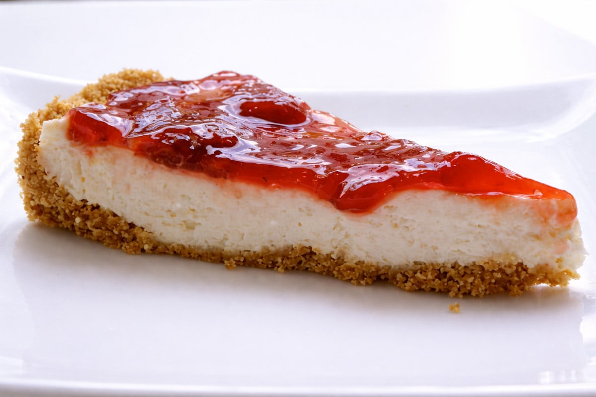 A slice of cheesecake with a crumbly graham cracker crust and a layer of bright red strawberry topping on a white plate.
