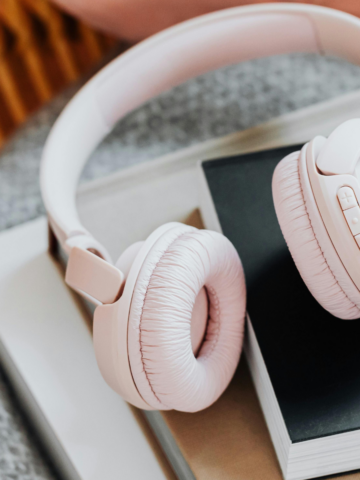 A pair of light pink noise canceling headphones for travel rests atop a stack of books with neutral-colored covers on a soft, patterned surface.