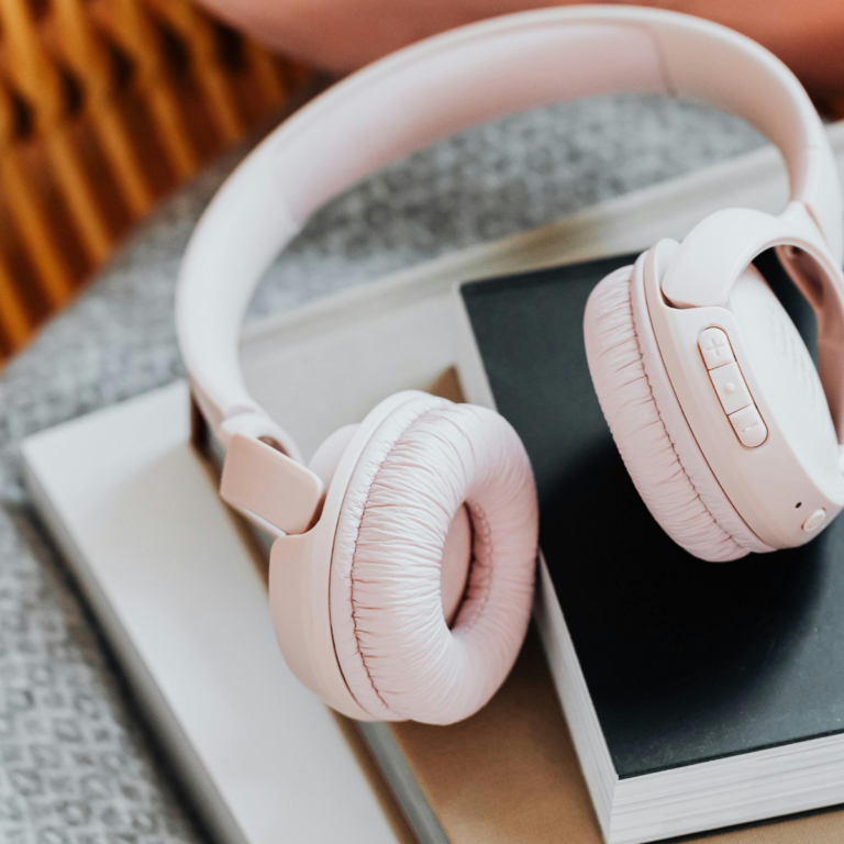 A pair of light pink noise canceling headphones for travel rests atop a stack of books with neutral-colored covers on a soft, patterned surface.