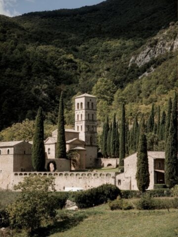 A stone abbey with a tall bell tower stands among tall cypress trees, surrounded by lush green hills and forests under a partly cloudy sky in Terni, Italy.