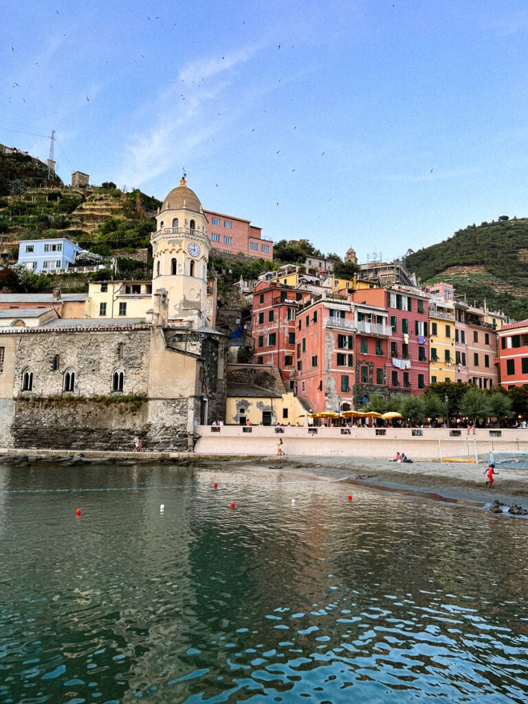 Colorful buildings and a church with a dome line Vernazza’s waterfront in Italy. The calm sea reflects the vibrant scene, while a sandy beach with a few people sits in the foreground and green hills rise behind.