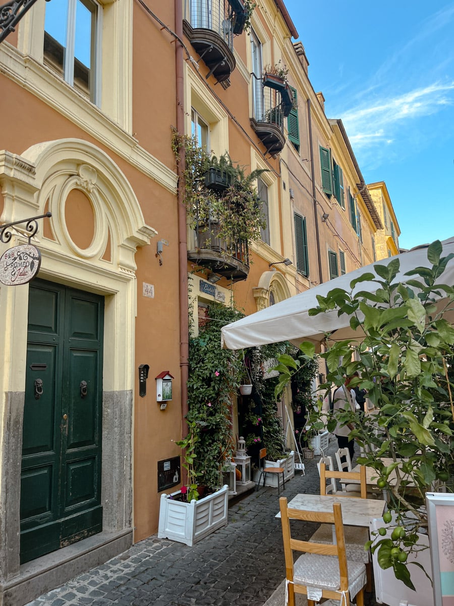 Narrow cobblestone street with colorful buildings, green doors, balconies with plants, and an outdoor café with wooden tables, chairs, and potted plants under a white canopy on a sunny day.