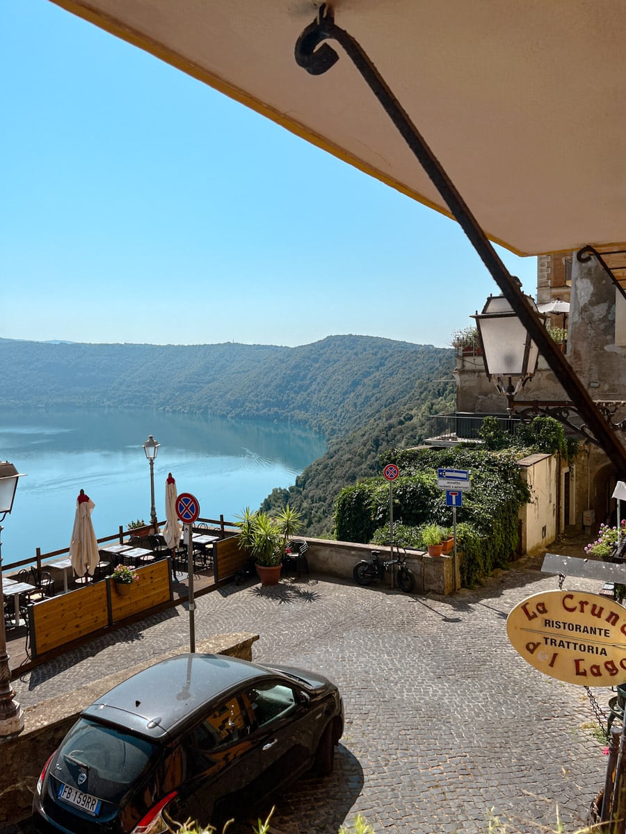 A scenic terrace overlooking a serene lake and green hills, with a cobblestone street, outdoor restaurant seating, potted plants, and a parked car in a charming hillside village.