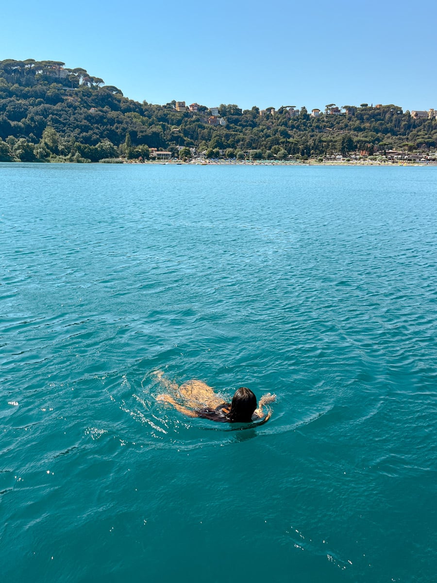 A person with long dark hair swims in clear blue water near a forested shoreline, with houses visible on the hillside under a clear sky.