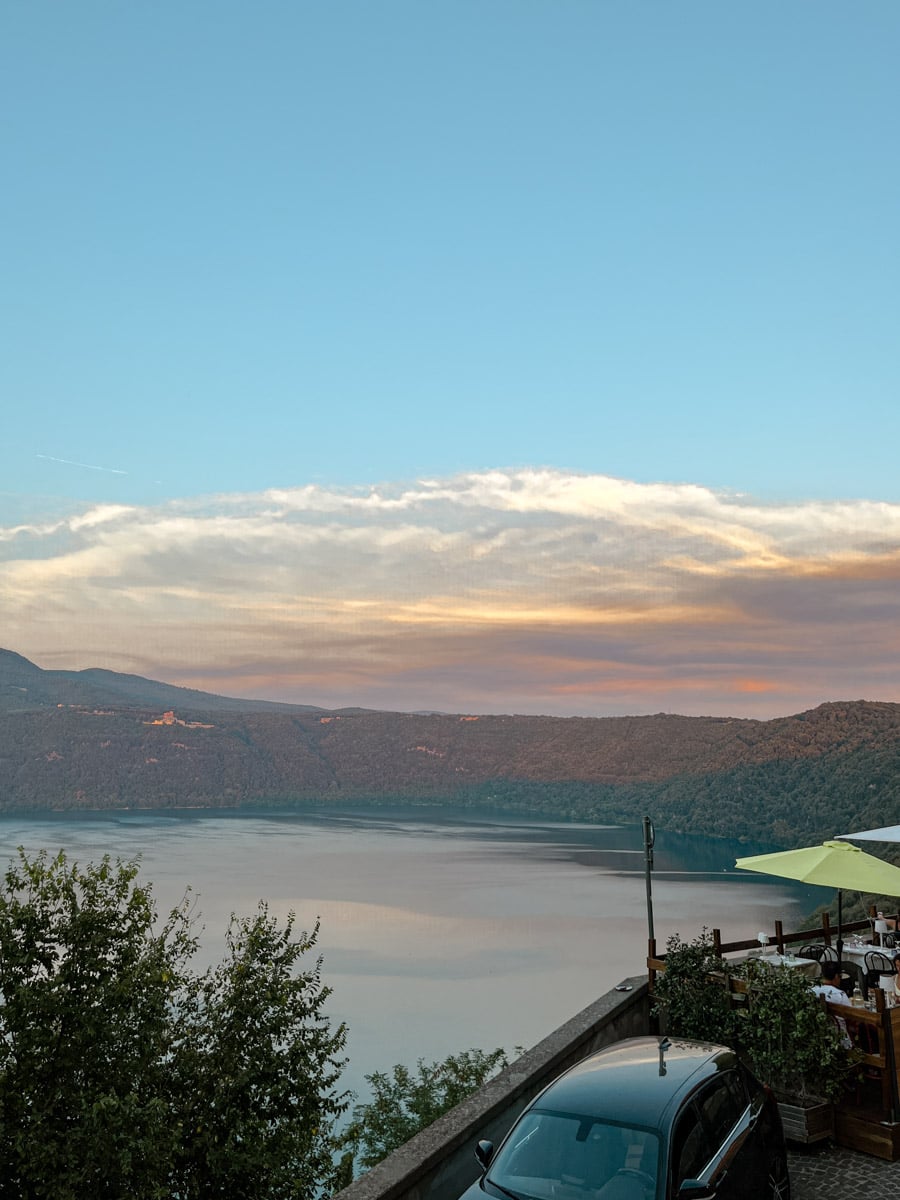 A serene lake surrounded by forested hills at sunset, with a parked car and outdoor dining area in the foreground under a clear sky with colorful clouds.
