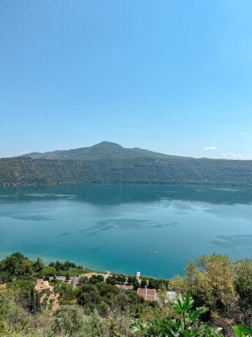 A clear blue lake is surrounded by green hills, with a few houses and trees in the foreground under a bright blue sky.
