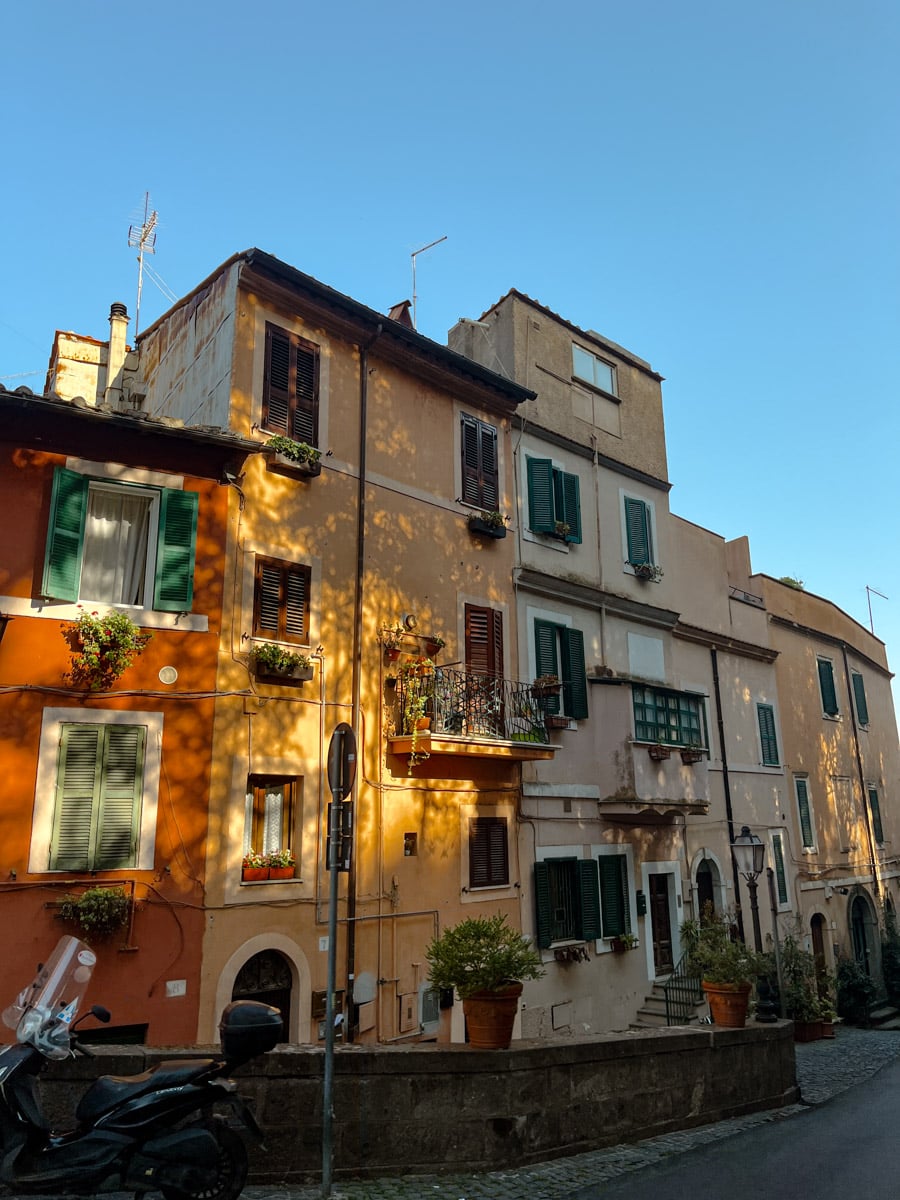 A row of colorful, sunlit buildings with green shutters and flower boxes line a narrow street under a clear blue sky. A motorcycle is parked in the foreground.