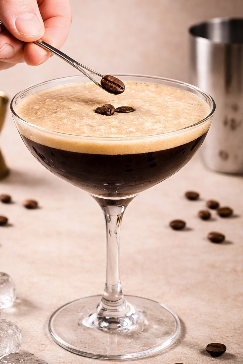 A hand holds a small spoon placing coffee beans on the frothy surface of an espresso martini in a coupe glass, with scattered coffee beans and a cocktail shaker in the background.