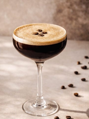 A coupe glass filled with a dark espresso martini, topped with a thick, frothy layer and garnished with three coffee beans. Scattered coffee beans are visible on the light-colored surface nearby.