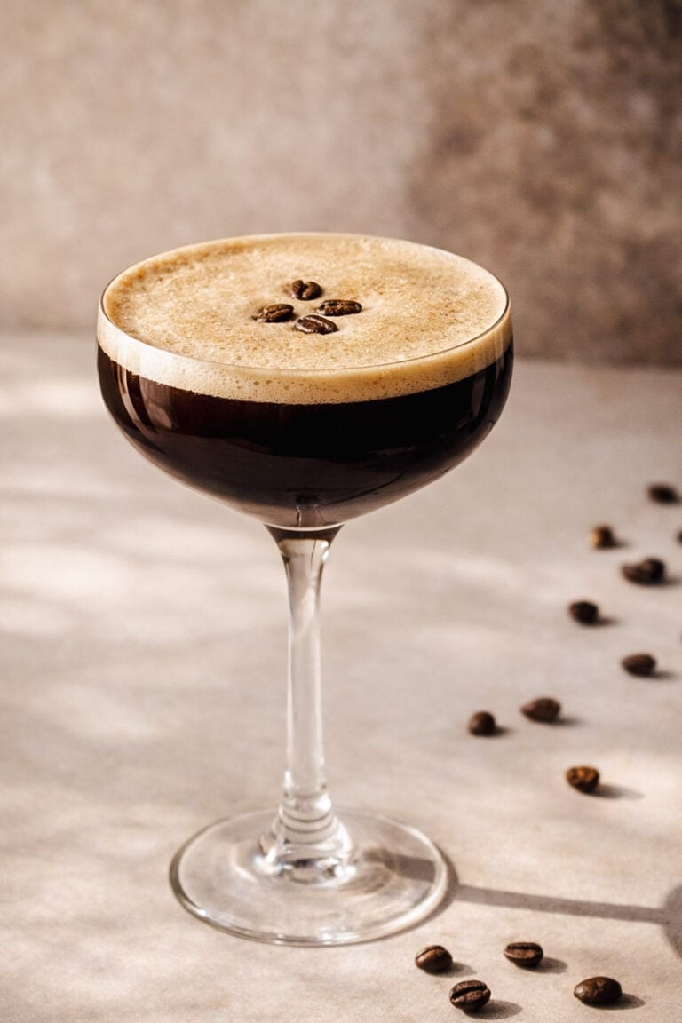 A coupe glass filled with a dark espresso martini, topped with a thick, frothy layer and garnished with three coffee beans. Scattered coffee beans are visible on the light-colored surface nearby.