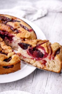 A close-up of a slice of plum cake being lifted from a round cake on a white plate. The cake has a golden crust, juicy plum pieces, and a soft, moist interior. The background is a light wooden surface.