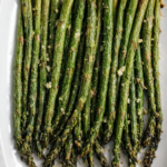 A close-up of roasted asparagus spears arranged neatly on a white oval plate, garnished with bits of garlic.