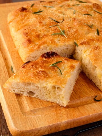 A rectangular piece of golden-brown focaccia bread topped with rosemary sits on a wooden cutting board, with one slice cut and slightly separated from the rest.