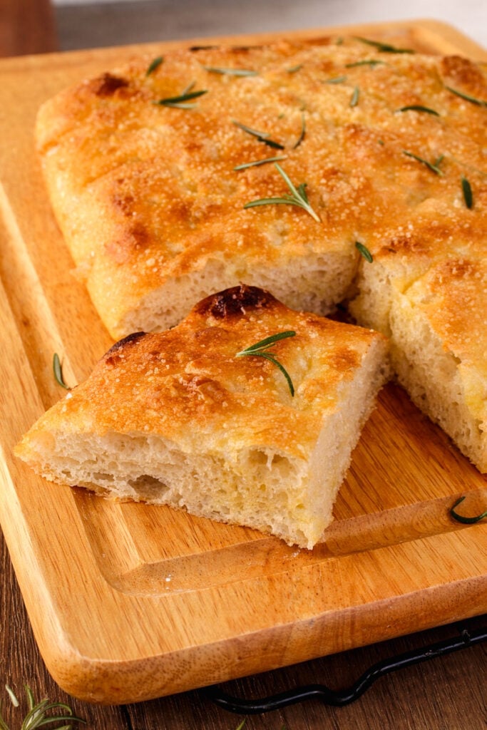 A rectangular piece of golden-brown focaccia bread topped with rosemary sits on a wooden cutting board, with one slice cut and slightly separated from the rest.