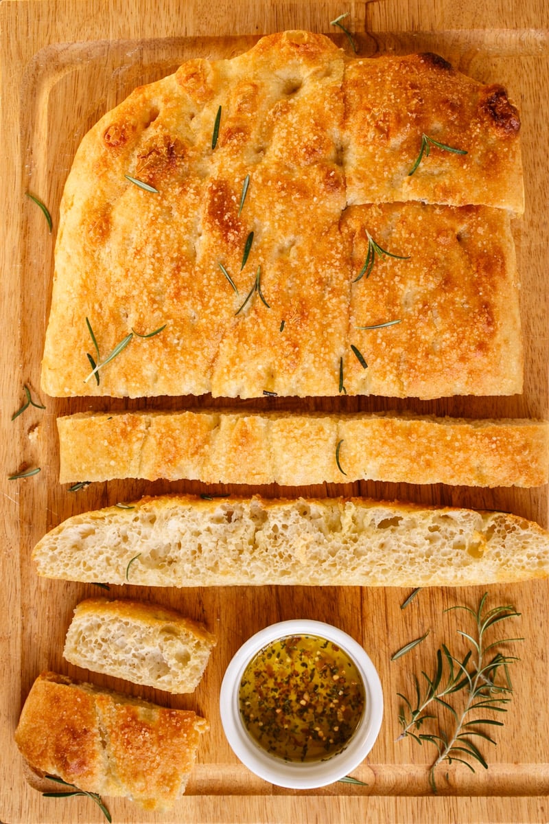 A loaf of sliced focaccia bread with rosemary sits on a wooden cutting board, accompanied by a small dish of olive oil and herbs for dipping.