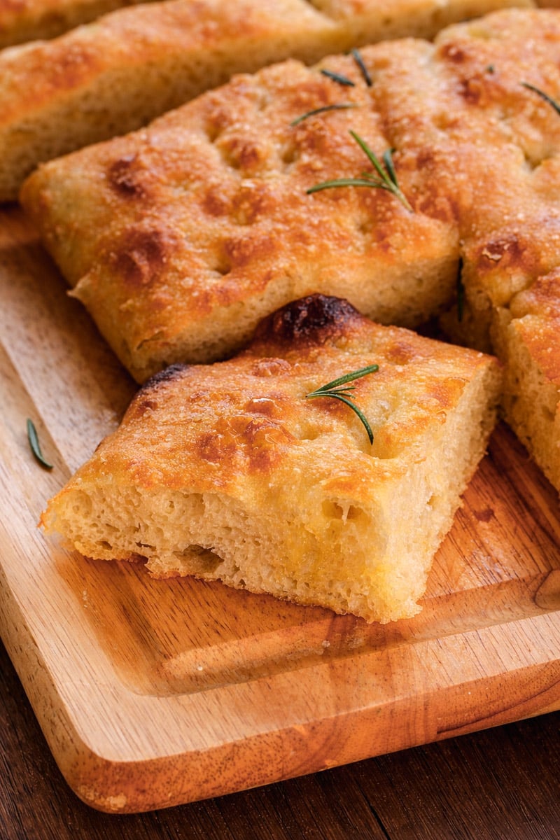Golden, airy slices of focaccia bread topped with rosemary rest on a wooden cutting board; one slice is slightly pulled out, showing its soft, fluffy interior.