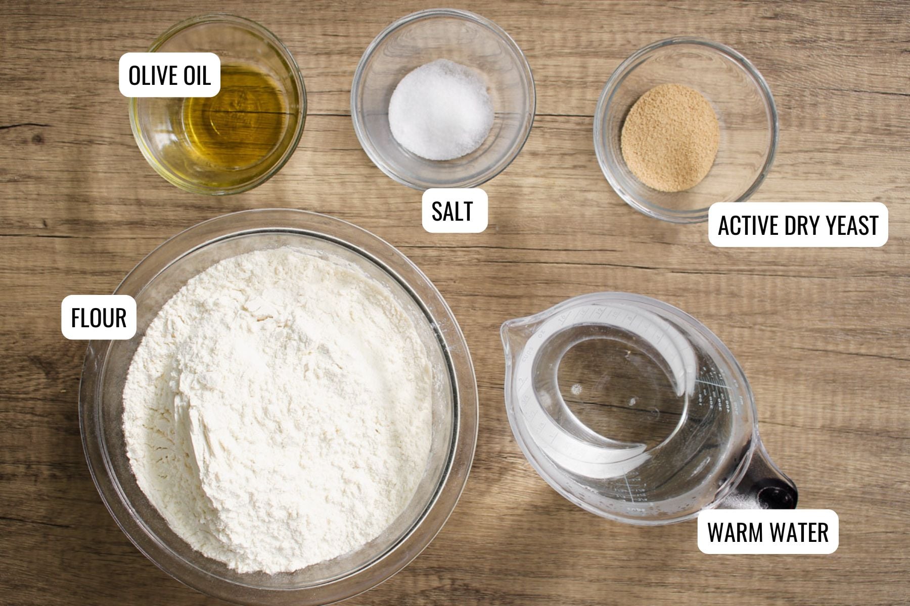 Five labeled bowls on a wooden surface containing olive oil, salt, active dry yeast, flour, and warm water, arranged for baking preparation.