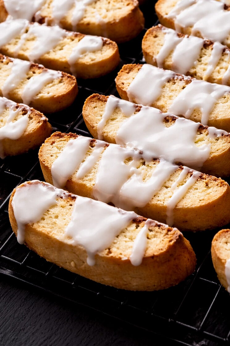 Rows of golden-brown Lemon Biscotti topped with white icing drizzle, cooling on a black wire rack. The biscotti are arranged neatly, and the glossy icing appears freshly applied.