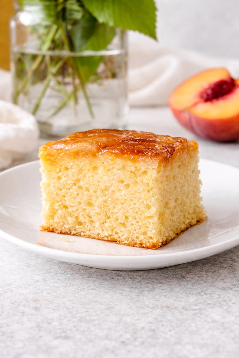 A square slice of moist Upside-Down Buttermilk Peach Cake with a shiny glaze sits on a white plate, accompanied by a peach half and a vase of green leaves in the blurred background.