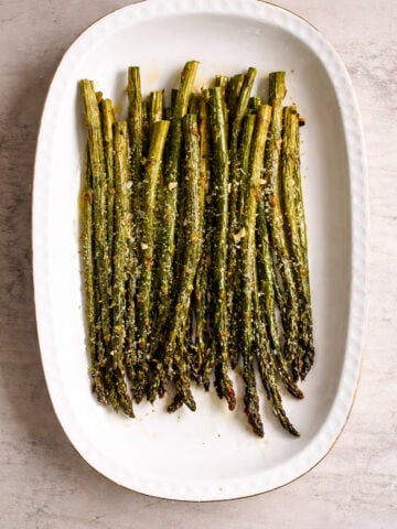 A white oval dish filled with roasted asparagus spears, sprinkled with grated cheese, on a light stone background.