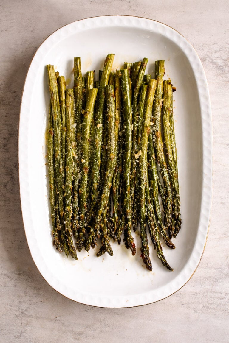 A white oval dish filled with roasted asparagus spears, sprinkled with grated cheese, on a light stone background.