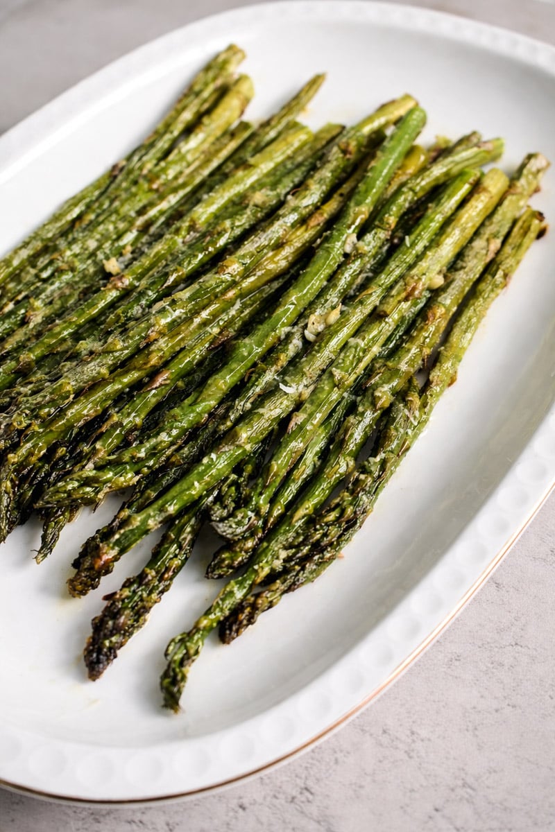 A white oval plate holds a neatly arranged pile of roasted asparagus spears, lightly charred and seasoned, on a light gray surface.