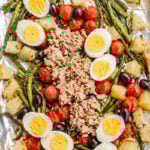 A baking sheet lined with foil holds roasted green beans, cherry tomatoes, potato cubes, olives, hard-boiled egg slices, and a mound of flaked tuna in the center.
