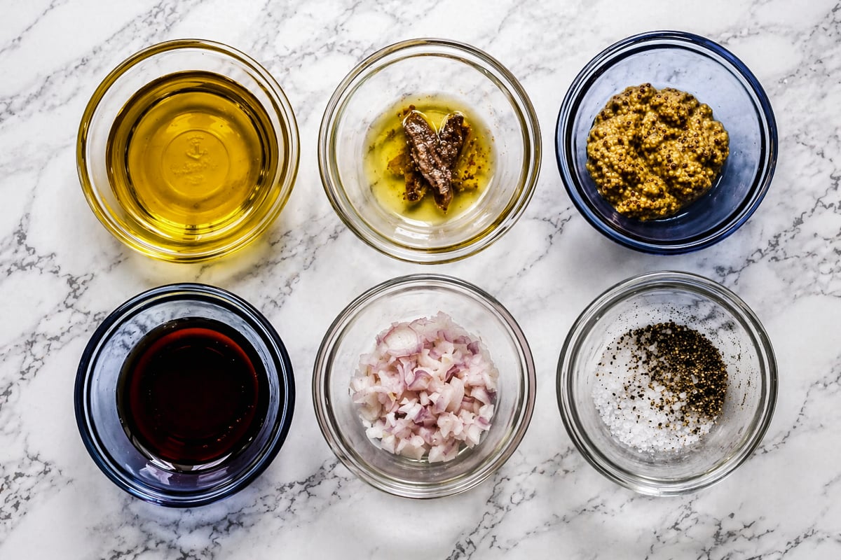 Six glass bowls on a marble surface containing olive oil, anchovies, whole grain mustard, red wine vinegar, chopped shallots, and a mixture of salt and black pepper.