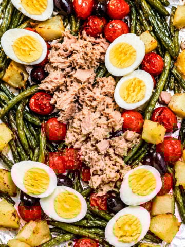 A sheet pan with green beans, roasted potatoes, cherry tomatoes, black olives, hard-boiled egg halves, and a mound of flaked tuna in the center.