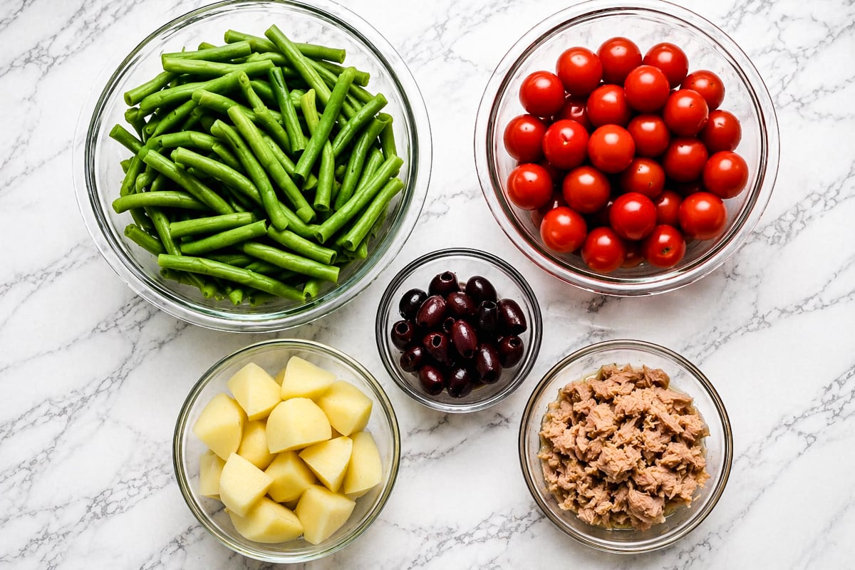Five glass bowls on a marble surface containing green beans, cherry tomatoes, black olives, cubed potatoes, and flaked tuna, all prepared and ready for cooking or assembling a dish.