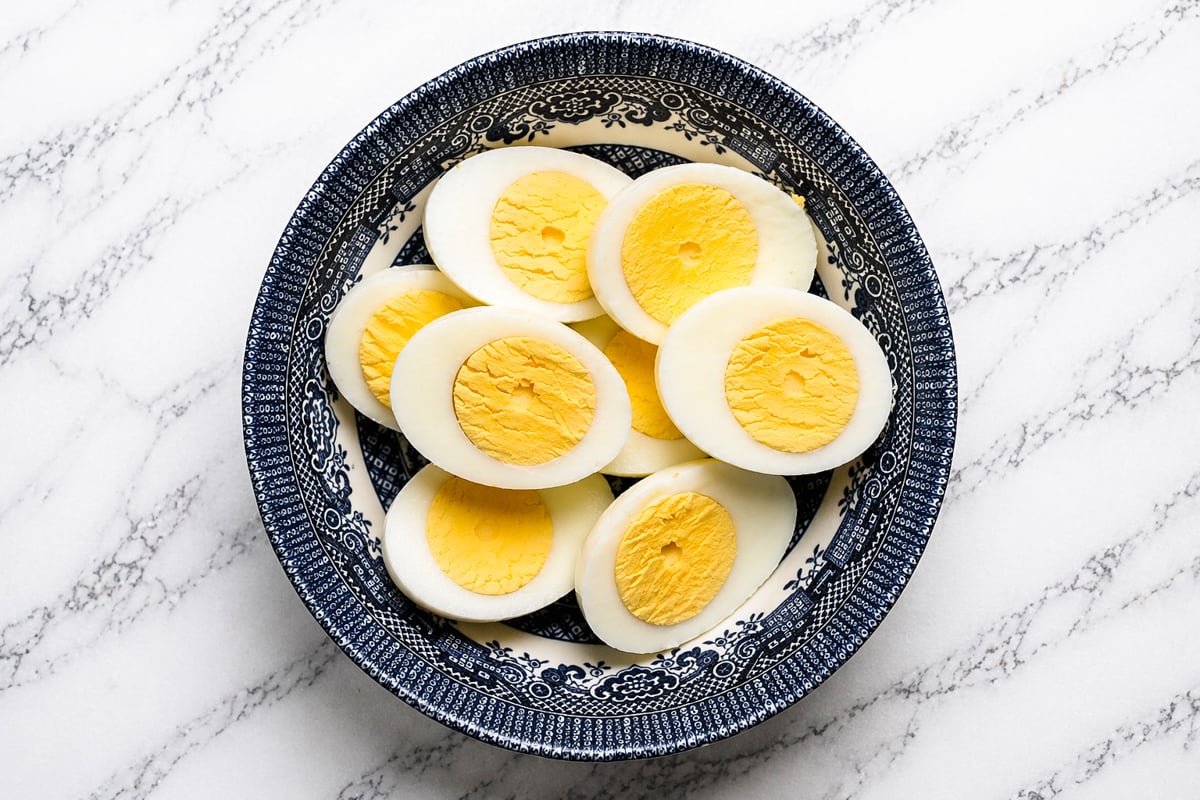 A blue and white patterned bowl filled with sliced hard-boiled eggs, showing yellow yolks and white edges, sits on a white marble surface.