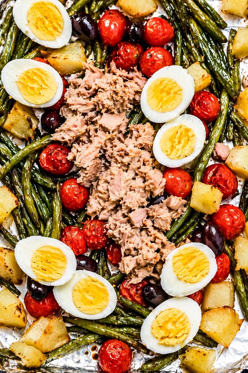 A sheet pan with green beans, roasted potatoes, cherry tomatoes, black olives, hard-boiled egg halves, and a mound of flaked tuna in the center.