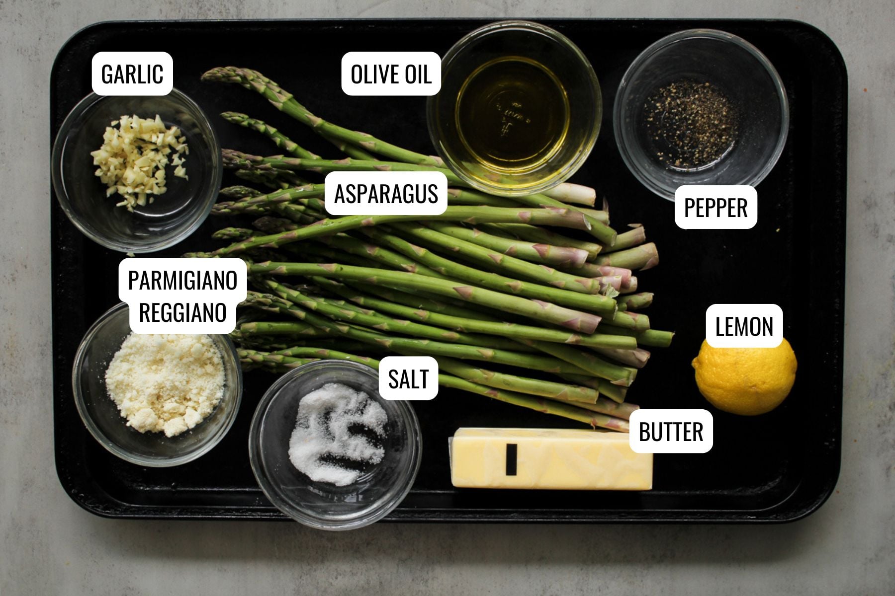 A tray with labeled ingredients: minced garlic, olive oil, ground pepper, asparagus spears, grated Parmigiano Reggiano, salt, half a lemon, and a stick of butter.