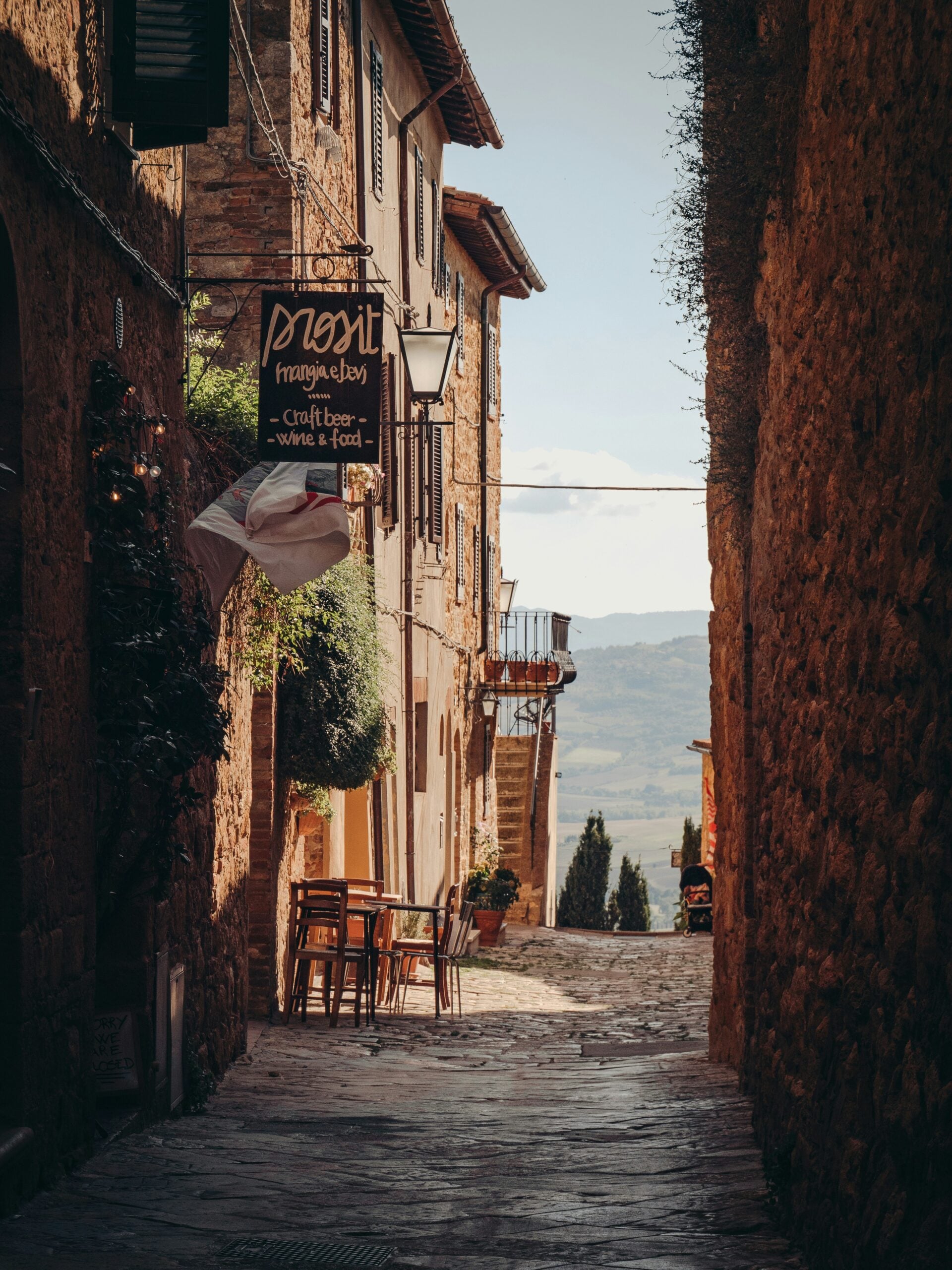 Narrow cobblestone street lined with rustic stone buildings, outdoor café tables set along the wall, a hanging sign reading L’Agona, and a distant scenic view of hills under a bright sky.