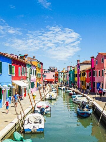 A canal in Burano lined with small boats and colorful houses on both sides under a bright blue sky; people walk along the waterfront in this vibrant, sunny neighborhood.