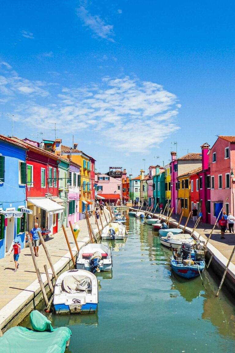A canal in Burano lined with small boats and colorful houses on both sides under a bright blue sky; people walk along the waterfront in this vibrant, sunny neighborhood.