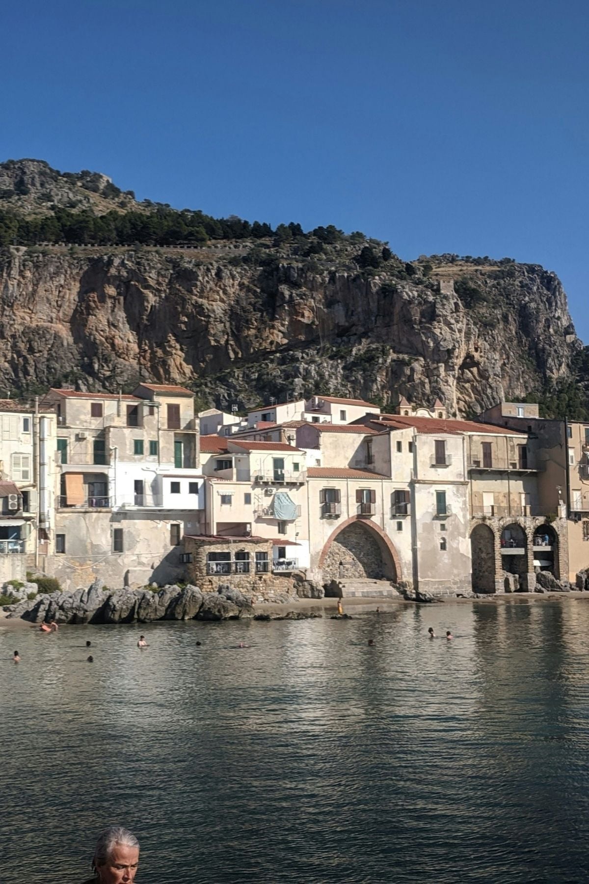 Buildings with light-colored walls sit by the edge of a calm body of water, with swimmers visible. Behind the buildings, there is a steep rocky cliff and blue sky above.