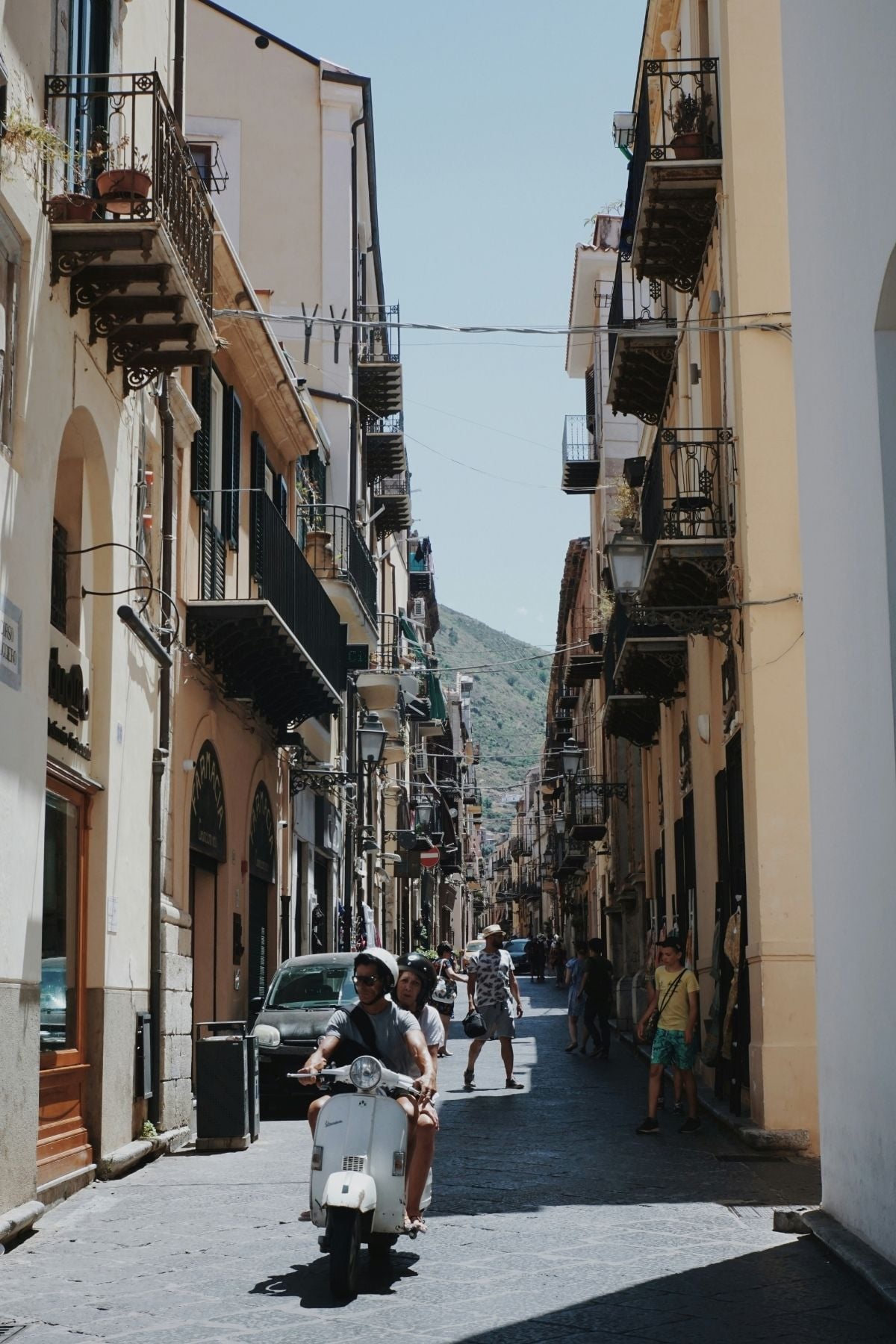 A narrow street in an Italian town, lined with tall buildings and balconies. People walk along the sides, and two people ride a white scooter down the center of the street. The background shows a green hillside.
