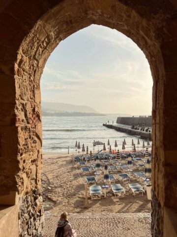A stone arch frames a view of a sandy beach with rows of closed umbrellas and empty lounge chairs. Waves gently hit the shore, and a person sits alone at the entrance, facing the sea under a partly cloudy sky.