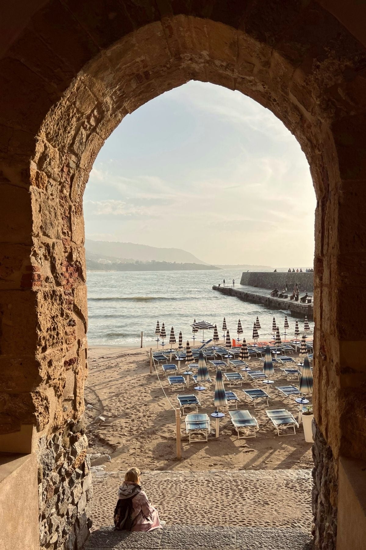 A stone arch frames a view of a sandy beach with rows of closed umbrellas and empty lounge chairs. Waves gently hit the shore, and a person sits alone at the entrance, facing the sea under a partly cloudy sky.