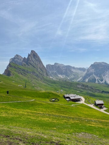 A scenic mountain landscape with rugged peaks, green grassy slopes, scattered buildings, and winding paths under a blue sky with soft clouds and sunlight streaming down.