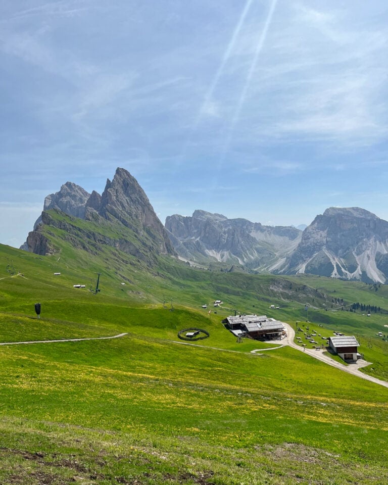 A scenic mountain landscape with rugged peaks, green grassy slopes, scattered buildings, and winding paths under a blue sky with soft clouds and sunlight streaming down.