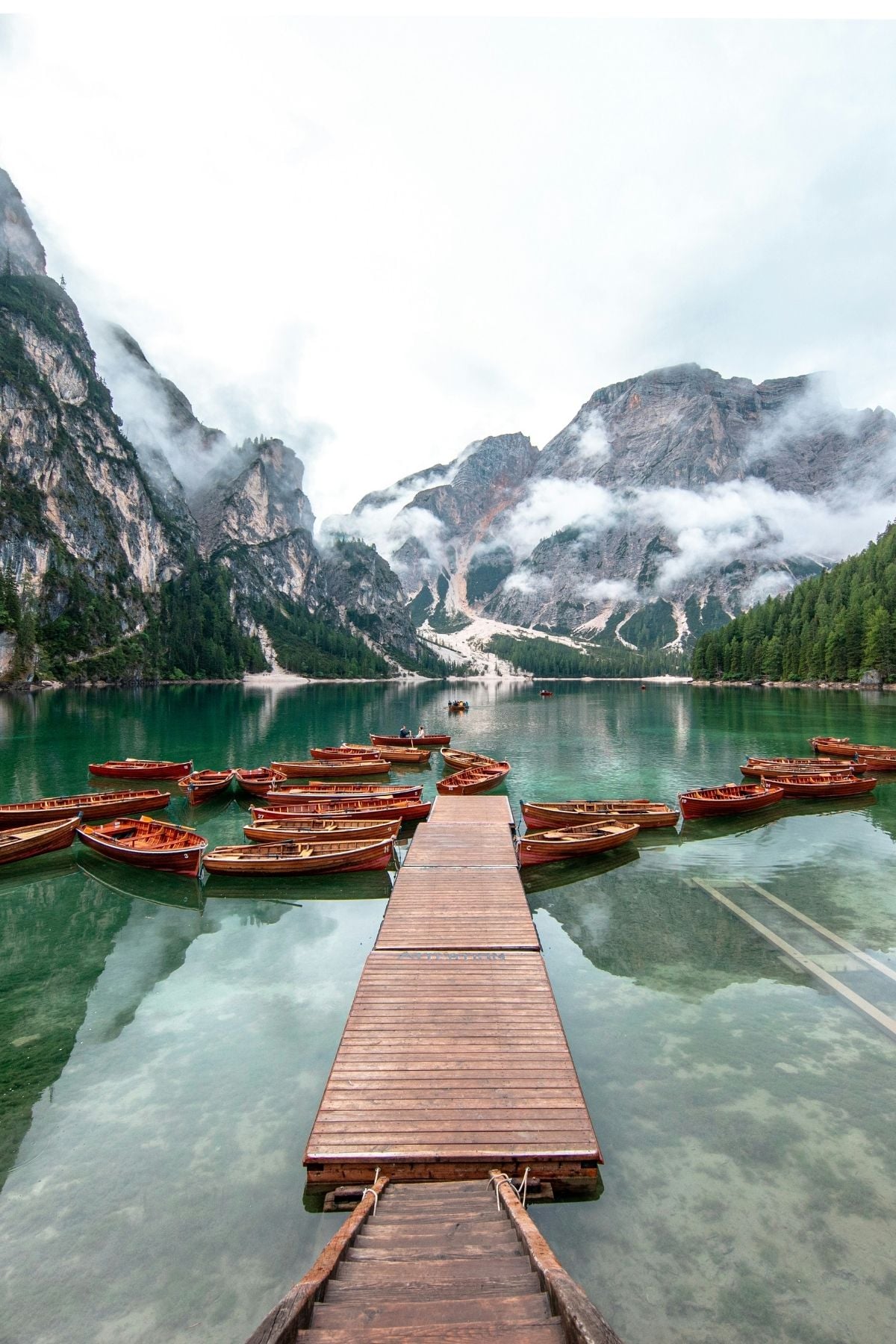 A wooden dock leads to small boats floating on a clear, turquoise lake surrounded by tall mountains and dense forest, with mist and clouds drifting among the peaks.