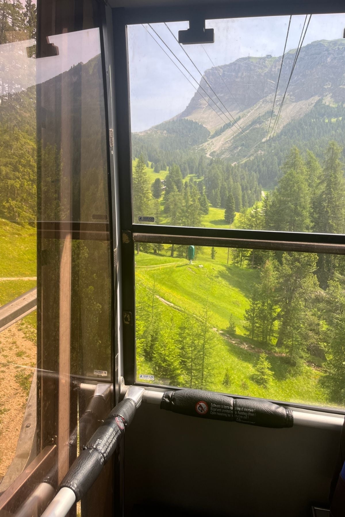 View from inside a cable car looking out over a lush, green mountain landscape with pine trees, grassy slopes, and a rocky peak in the background under a partly cloudy sky.