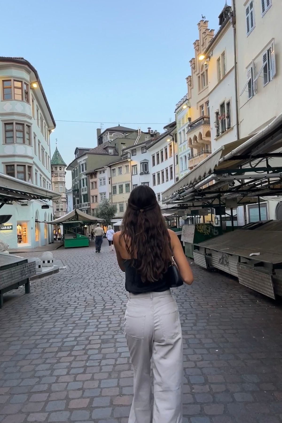 A woman with long brown hair, wearing a black top and white pants, walks down a cobblestone street lined with European-style buildings and closed market stalls at dusk.